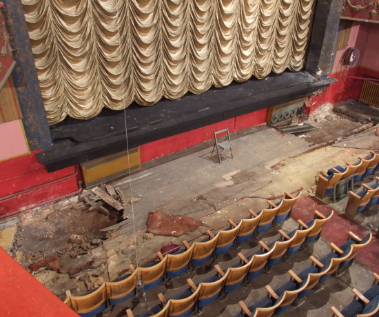 Auditorium of derelict Hyde Theatre Royal with damaged area in front of the stage, rows of seats and draped curtain in place.