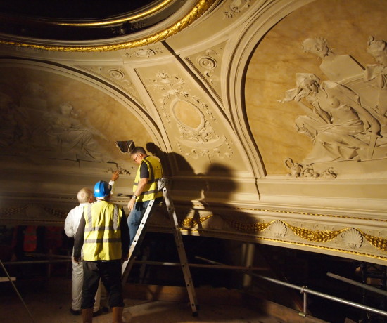 Ornate plasterwork of a domed theatre ceiling with three men in hi-vis vests inspecting it.
