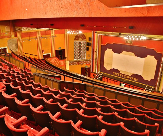 Orange, red and gold accented Art Deco Tameside Hippodrome auditorium, from the back of the steep-raked, red seated balcony down to the safety curtain with SAFETY CURTAIN written in gold letering on a being and maroon background