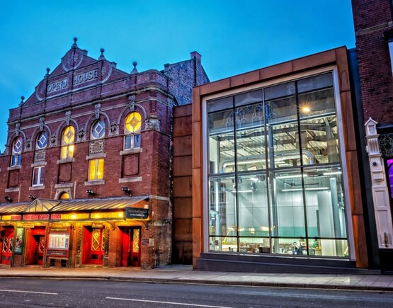 Exterior of Theatre Royal Wakefield viewed from the street at dusk. A grand historic brick building with round stained glass windows next to a modern glass-fronted extension building.