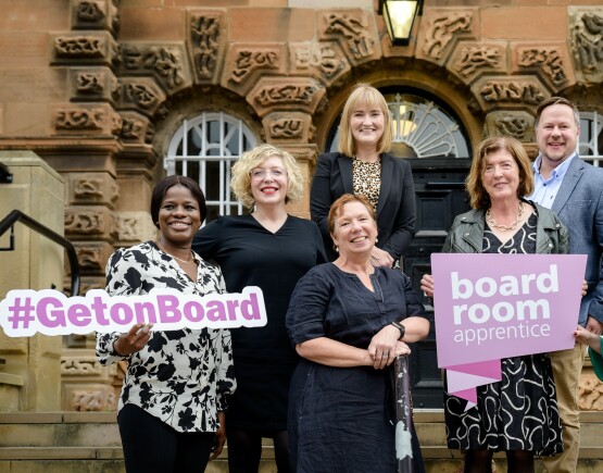 Group of people stood outside a stone building holding signs saying Boardroom Apprentice and #GetOnBoard