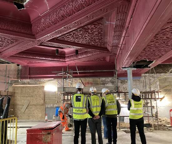 Group of people in hard hats and yellow safety jackets in a historic theatre auditorium with pink plaster ceiling that is being renovated.
