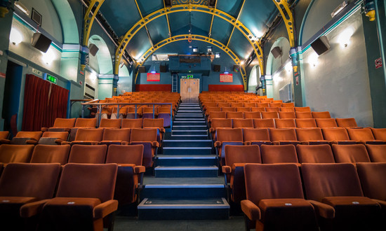 A view from the stage across the raked orange velour colour seats at Wallingford Corn Exchange with its blue central aisle stairs.