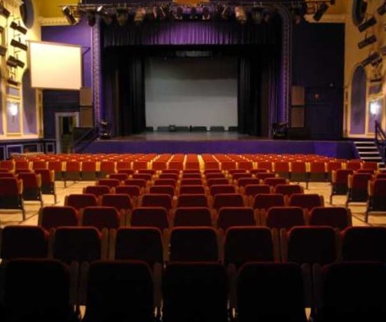 The auditorium of Shanklin Theatre on the Isle of Wight, from about 2016. It is a view from the back of the stalls towards the stage. The seats are red and stage curtains a deep purple.