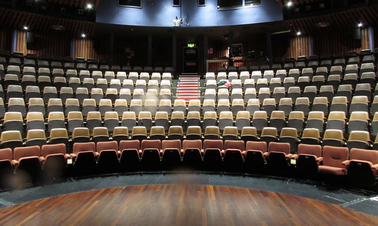The auditorium of Theatr Ardudwy with its sweep of steeply raked seating focused on an open stage with small curved thrust. Seats are light rust and faded mustard with backing in black