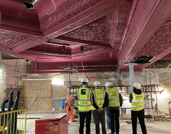 Group of people in hard hats and yellow safety jackets in a historic theatre auditorium with pink plaster ceiling that is being renovated.