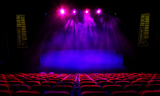 Lit auditorium with red seating in a single rake and a blank stage lit with purple spots.