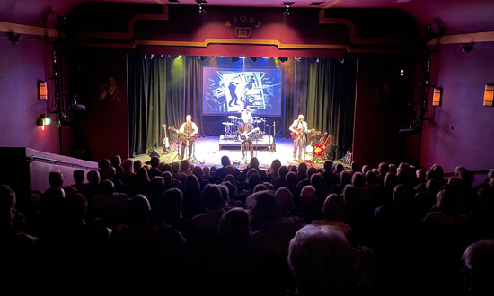 Performers on stage in a small theatre viewed across a packed audience.