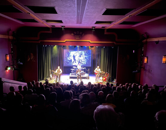 Performers on stage in a small theatre viewed across a packed audience.